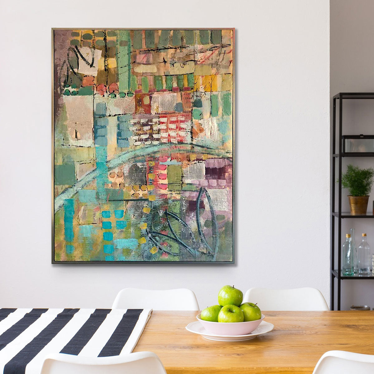 An abstract encaustic painting framed in a gold floater frame, hung on a white wall. Below the painting is a wooden table with a blue and white striped table runner and a bowl of green apples.