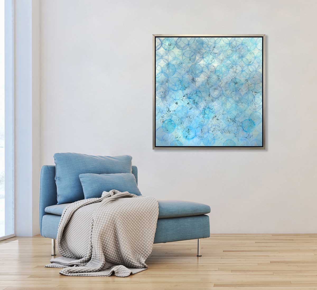 A blue and silver abstract circle print in a silver floater frame hangs on a white wall above a blue armless chair with a white throw blanket.