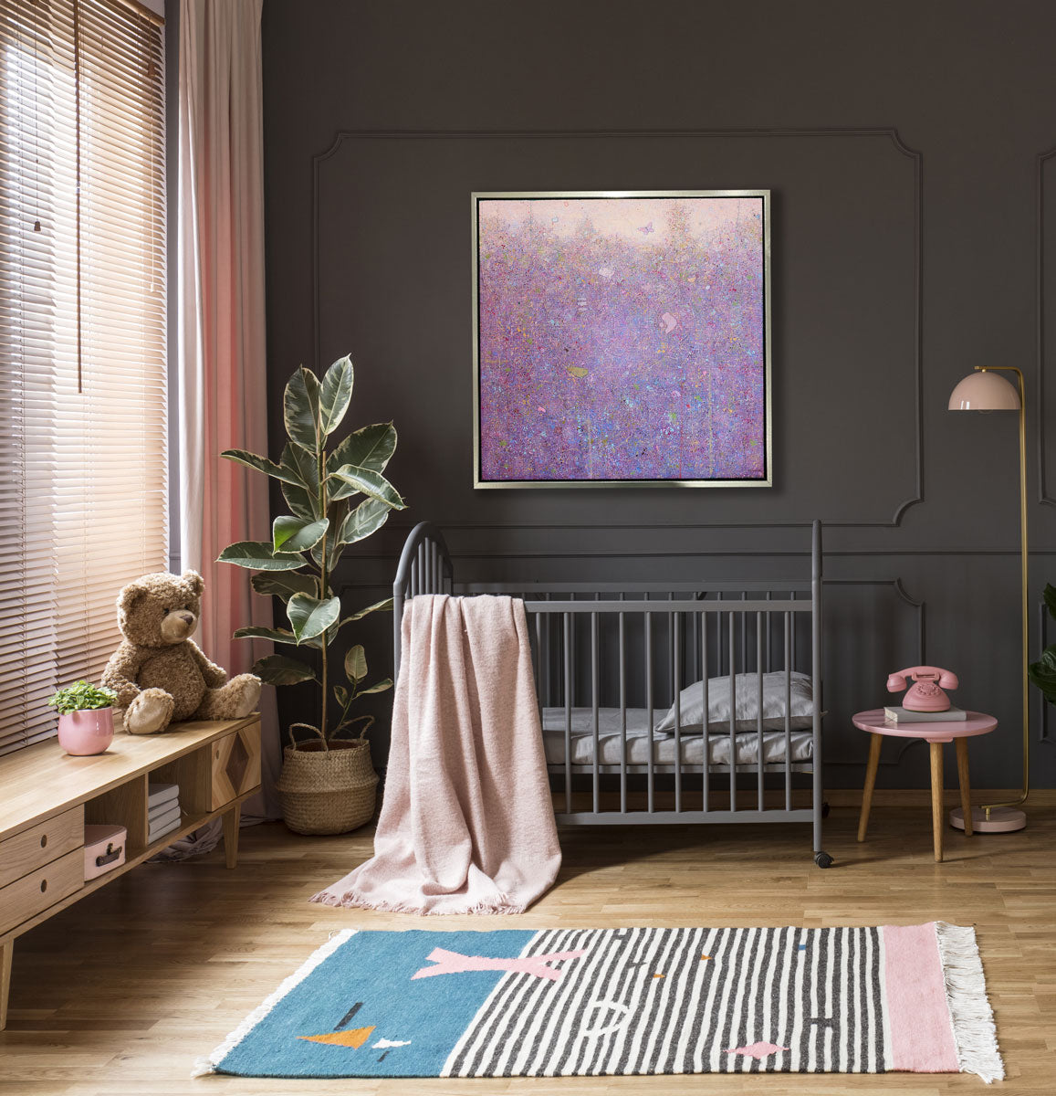 A pink abstract landscape print in a silver floater frame hangs on a dark grey wall above a grey crib with a blush colored blanket draped over the side, a potted floor plant and a low natural wood cabinet with a teddy bear.