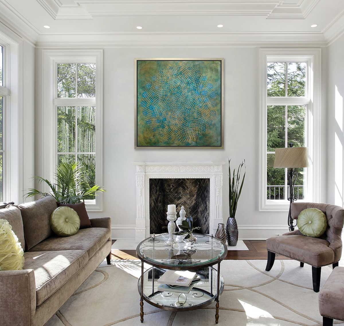 A green, blue and gold abstract overlapping circle print in a silver floater frame hangs on a white wall above a fireplace, a beige sofa with light green pillows, a light beige matching chair and a oval glass and iron coffee table.