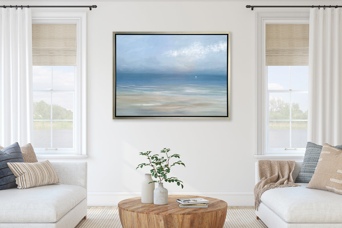 A light blue, white and beige abstract seascape print in a silver floater frame hangs on a white wall in between two large windows and above two sections of a white sofa with neutral pillows and a rounded wood coffee table with a stack of books and two vases with greenery stems.