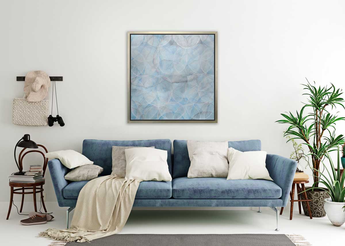 A light blue abstract overlapping circle print in a silver floater frame hangs on a white wall above a blue sofa with white pillows, two potted floor plants and a dark wood chair with books and a desk lamp.