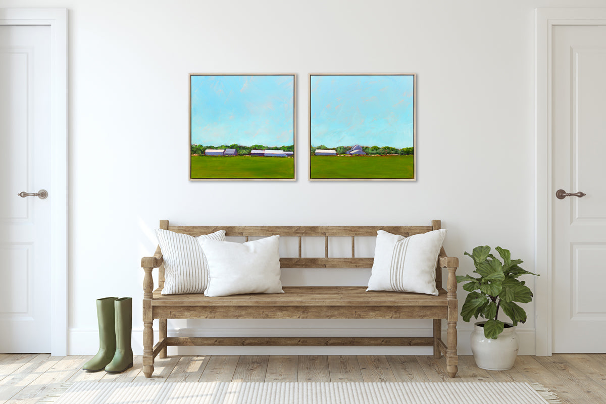 Two blue and green landscape prints in white oak wood floater frames hang side by side on a white wall above a natural wood bench with white pillows, a potted plant and a pair of green rubber boots.