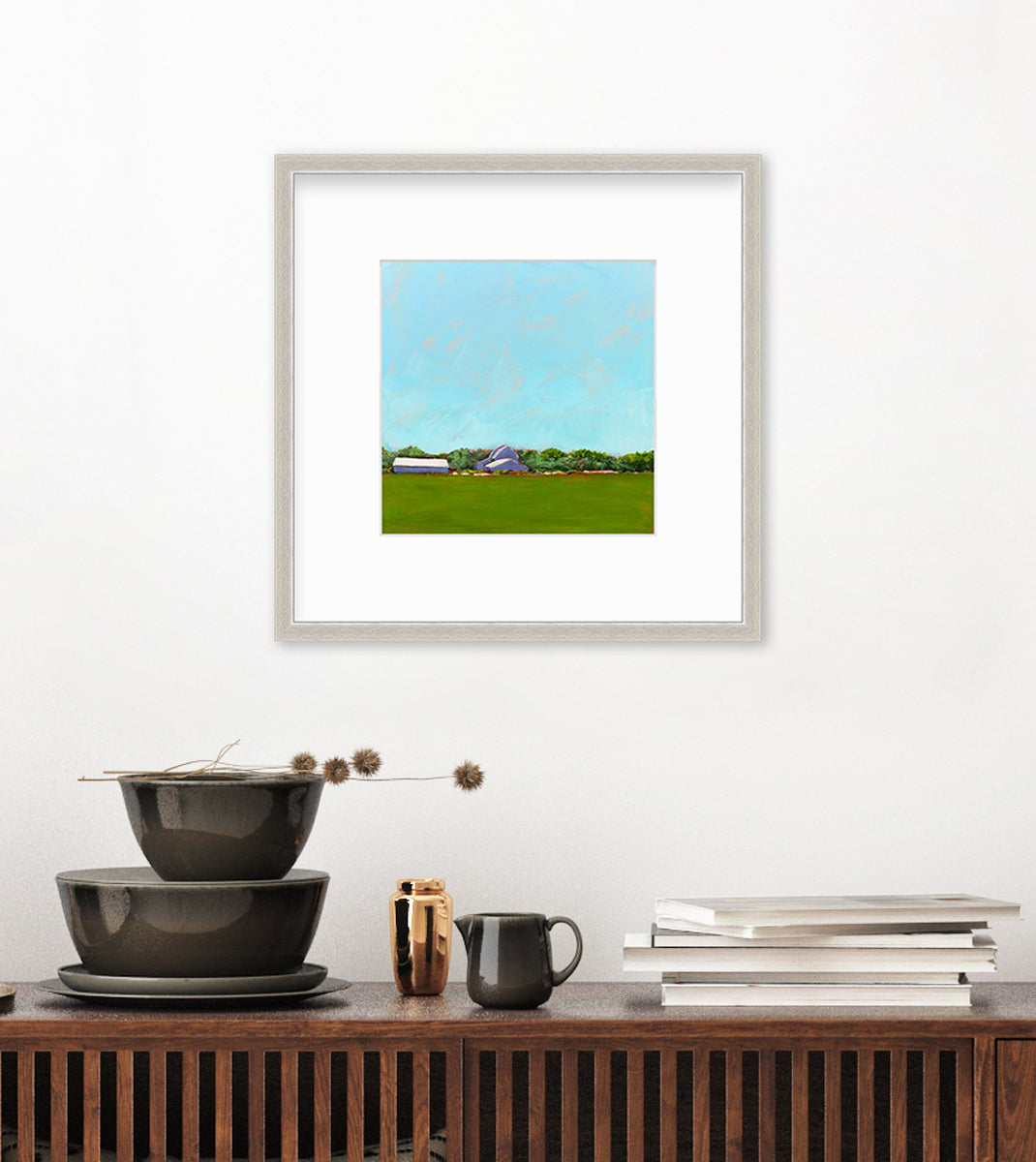 A blue, green, lavender and white landscape print in a silver frame with a mat hangs on a white wall above a dark wood surface with a stack of brown bowls and a stack of books.
