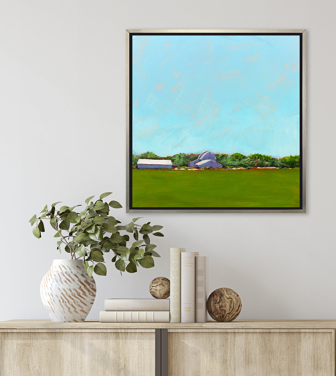 A blue, green, lavender and white landscape print in a silver floater frame hangs on a white wall above a natural wood cabinet with a stack of books, two wood sphere decorations and a white vase with greenery stems.