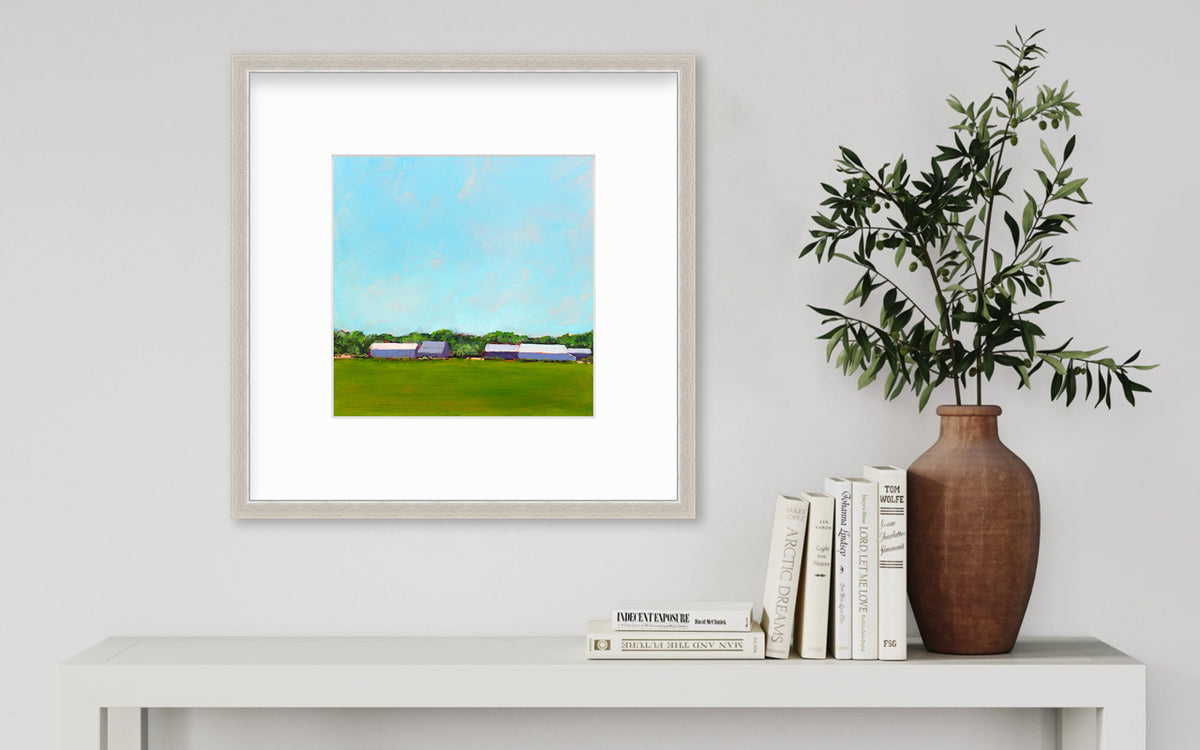 A blue, green, lavender and white landscape print in a silver frame with a mat hangs on a white wall above a white console table with a stack of books and a terracotta vase with greenery stems.