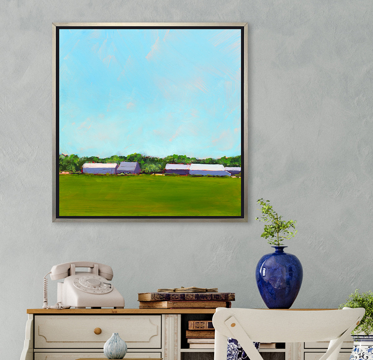A blue, green, lavender and white landscape print in a silver floater frame hangs on a light grey wall above a white desk with a white rotary phone, a stack of books and a small blue vase with a greenery stem.