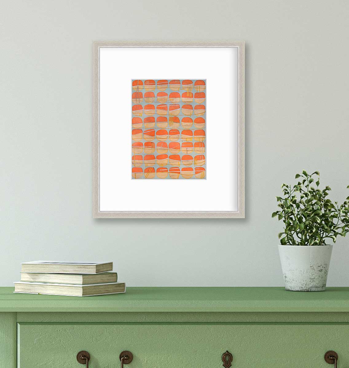 A orange, white and light blue abstract circle print in a silver frame with a mat hangs on a white wall above a green console table with a stack of books and a potted plant.