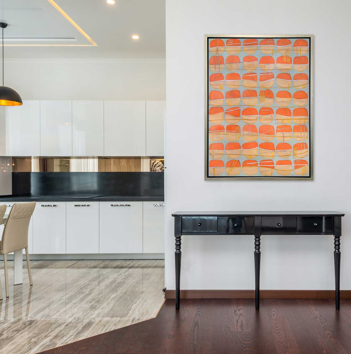 A orange, white and light blue abstract circle print in a silver floater frame hangs on a white wall above a black console table next to an opening of a kitchen.