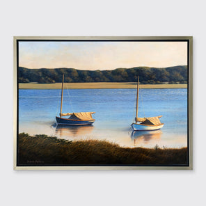 A contemporary realist print of two anchored boats wading in the water in a silver floater frame hangs on a white wall.