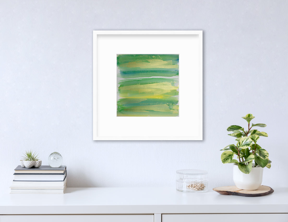 A green, blue and gold abstract print in a white frame with a mat hangs on a white wall above a white surface with a stack of books, a round clear container with gold accessories and a potted plant.