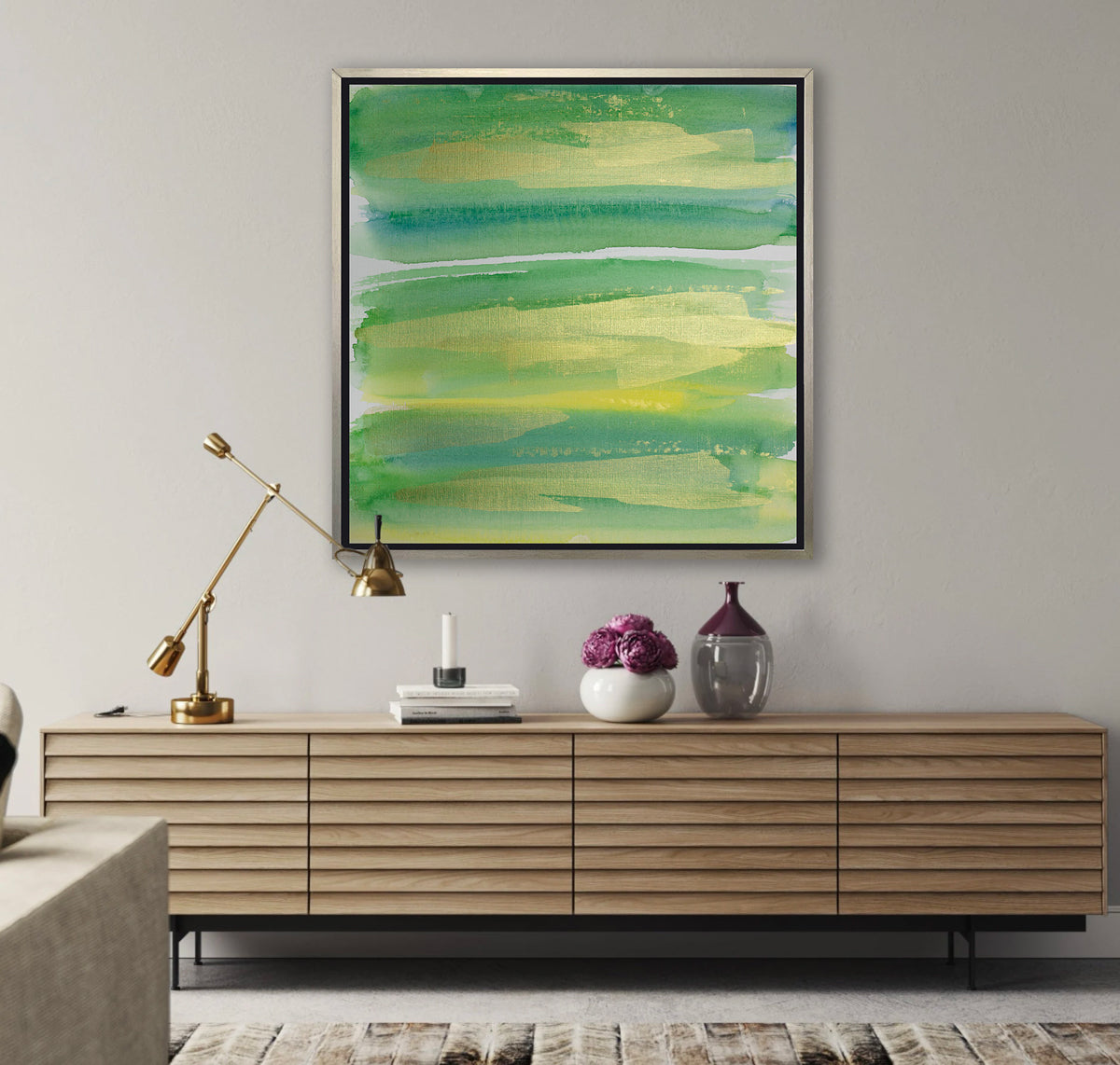 A green, blue and gold abstract print in a silver floater frame hangs on a white wall above a long wood console cabinet with a movable gold desk lamp, a stack of books and a white vase with purple flowers.