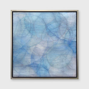 A multiple shades of blue overlapping circles with an iridescence in a silver floater frame hangs on a white wall.