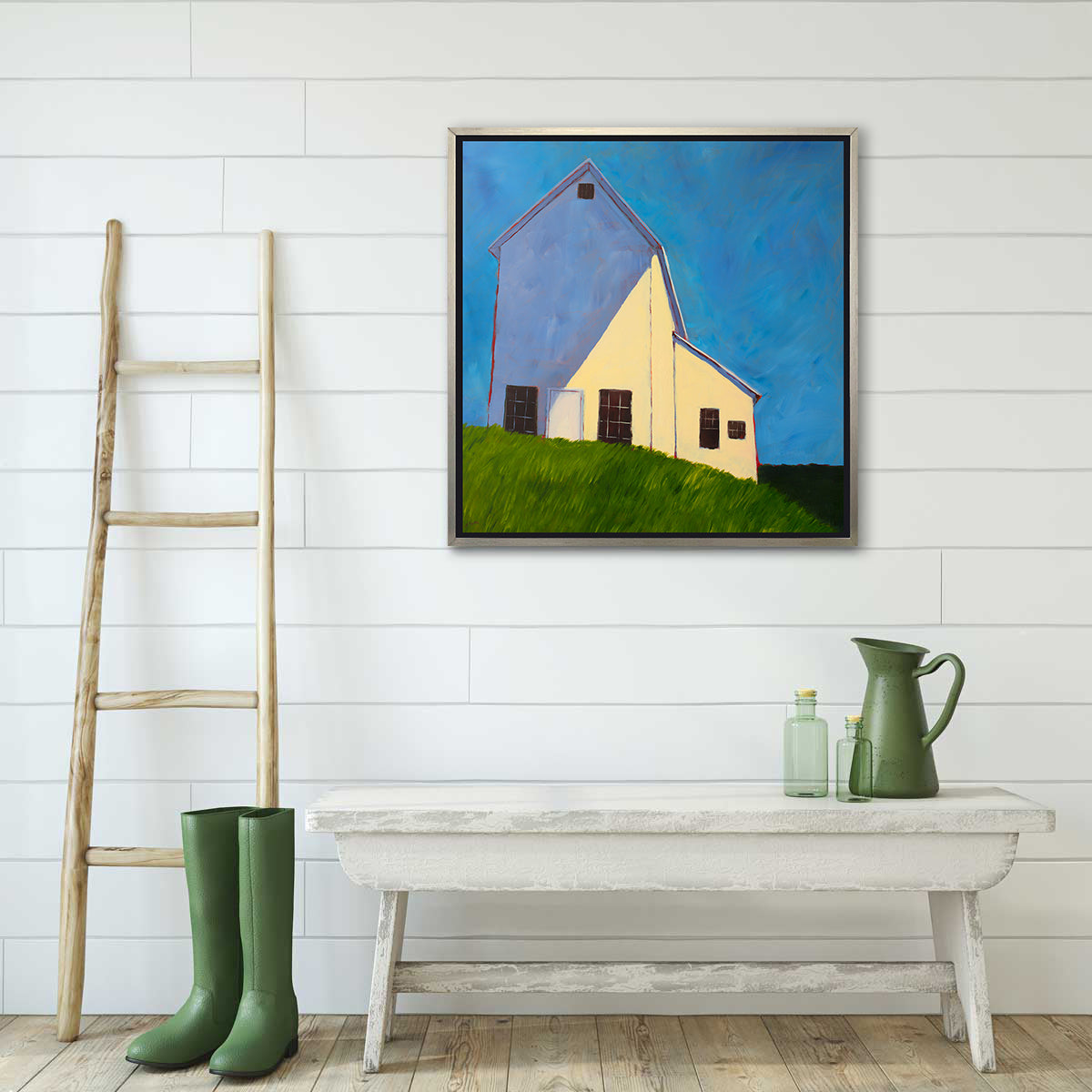 A blue, beige, lavender and green contemporary barn print in a silver floater frame hangs on a white sided wall above a whitewashed bench with a green pitcher, small green bottles and a pair of green rain boots in front of a ladder leaning on the wall.