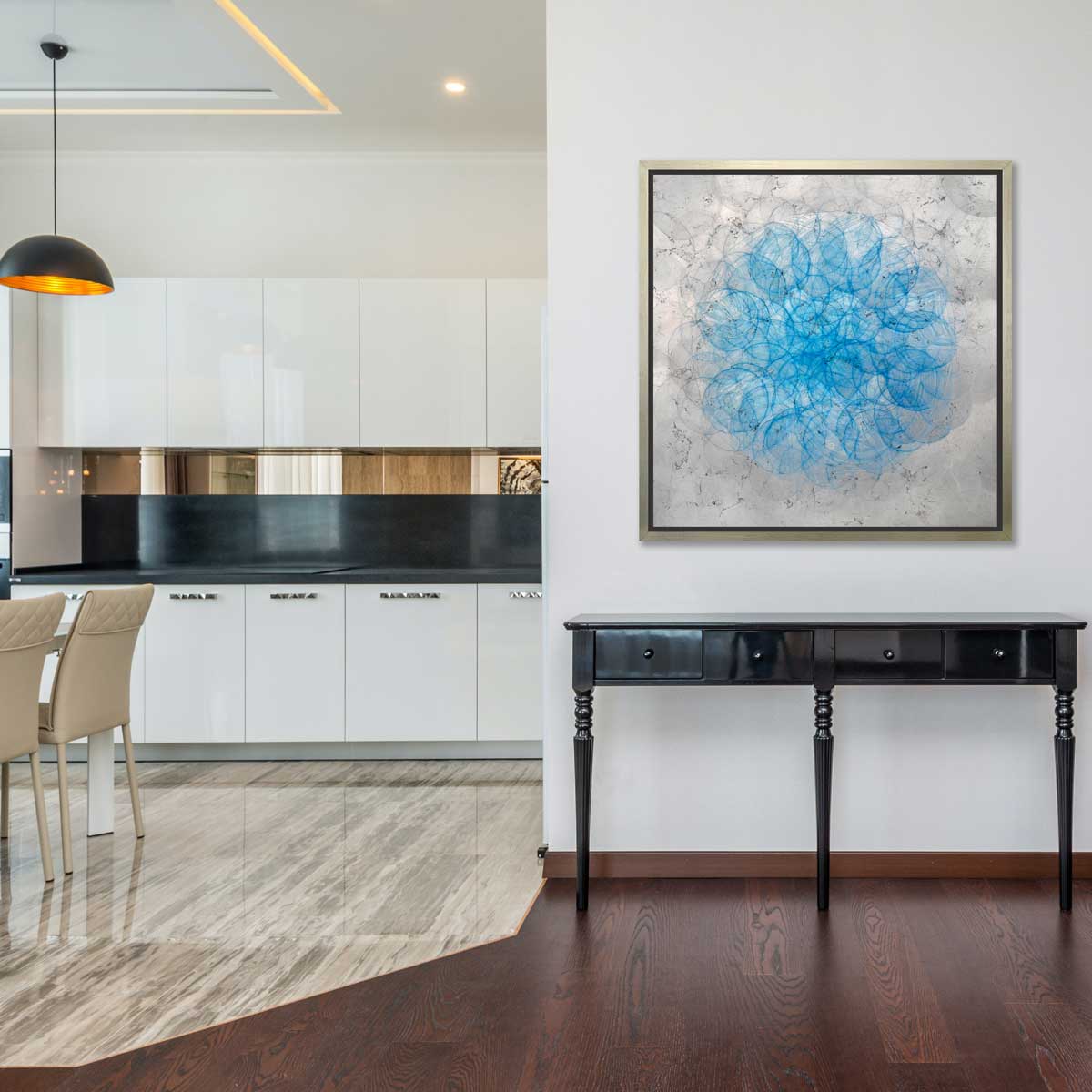 A silver, grey and blue abstract overlapping circle print in a silver floater frame hangs on a white wall above a black console table and next to an opening to a kitchen with black countertops, white cabinets and two beige chairs.