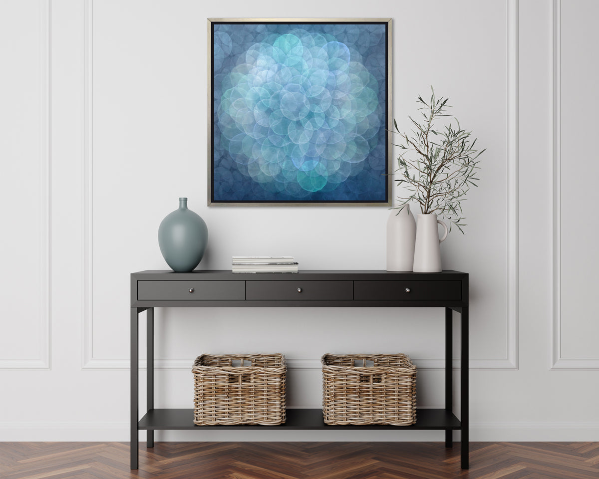 A blue, teal, light lavender and white abstract geometric print in a silver floater frame hangs on a white wall above a black console table with a muted teal vase, two greenery filled white vases and two woven baskets.