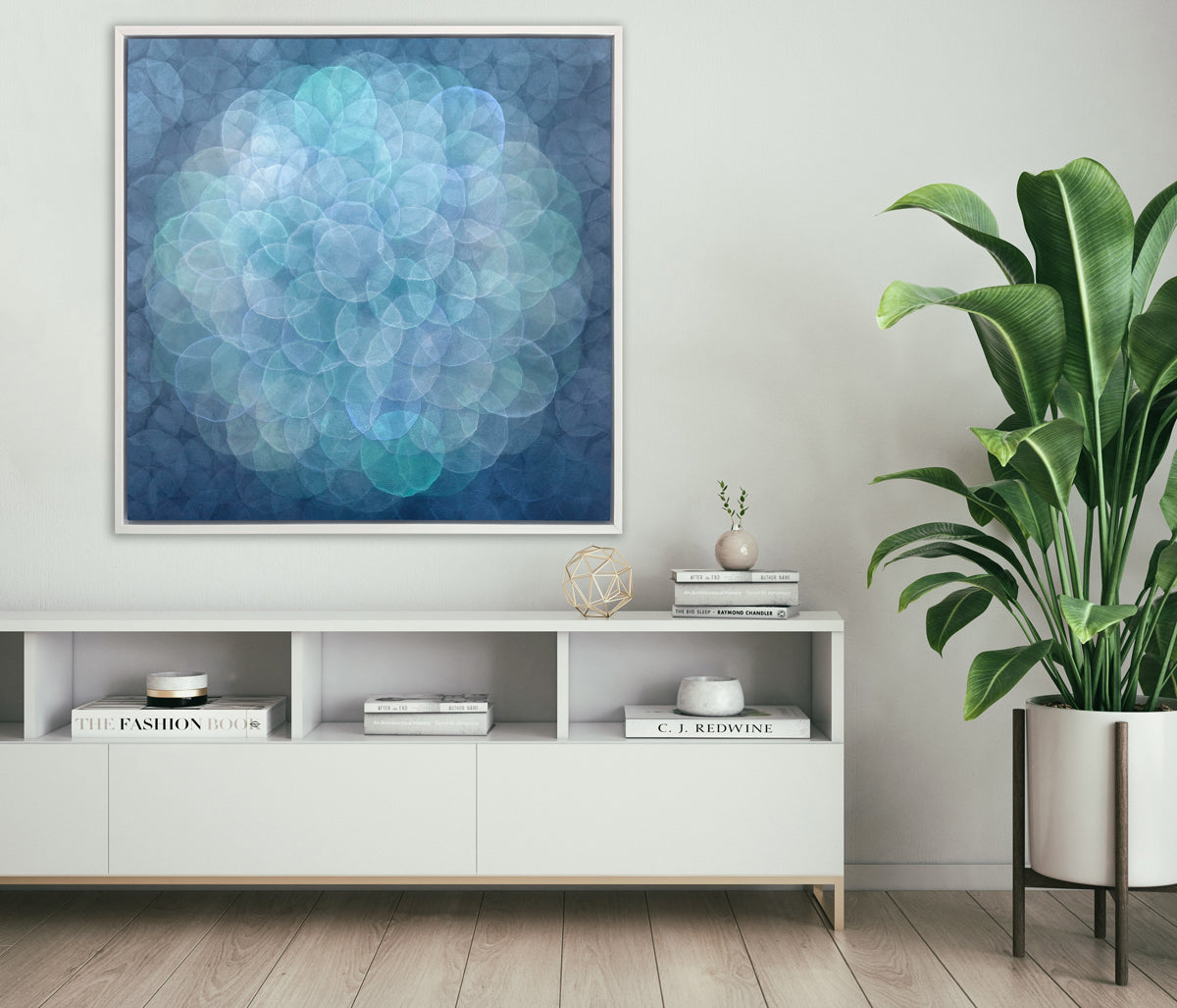 A blue, teal, light lavender and white abstract geometric print in a white floater frame hangs on a white wall above a white credenza with stacks of books, a gold wire sphere decoration and a tall potted floor plant to the right.
