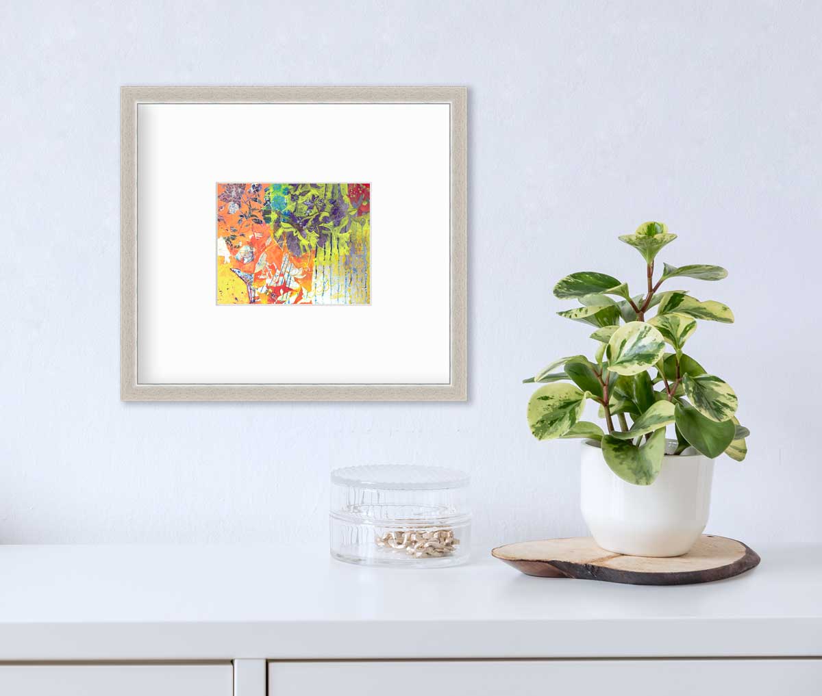 A multicolored abstract print in a silver frame with a mat hangs on a white wall above a white surface with a clear container holding brushed gold baubles, a tree trivet and potted plant.