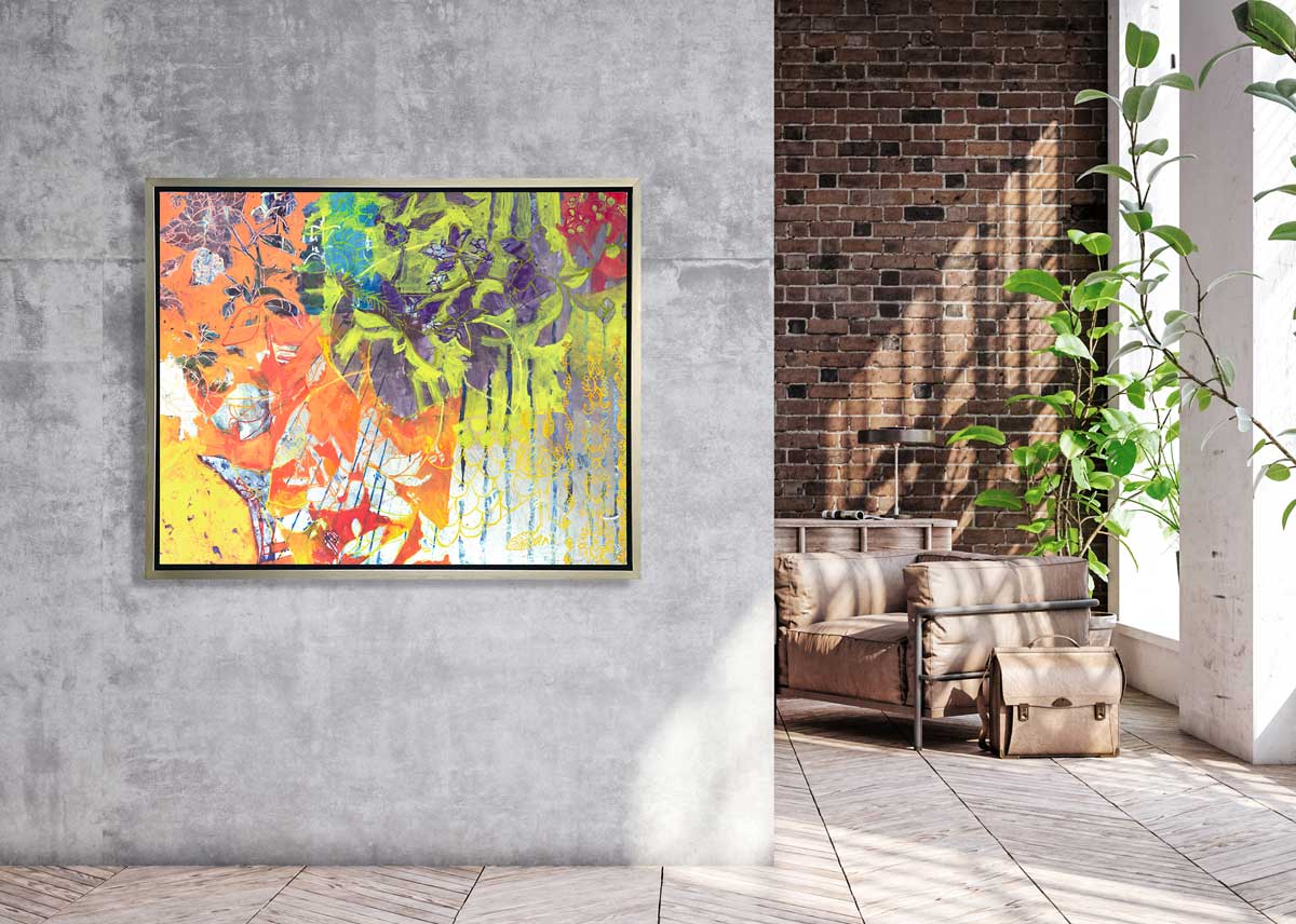 A multicolored abstract print in a silver floater frame hangs on a concrete grey wall next to an opening to a brick wall, an overstuffed brown chair, a leather briefcase and large windows with a potted floor plant.