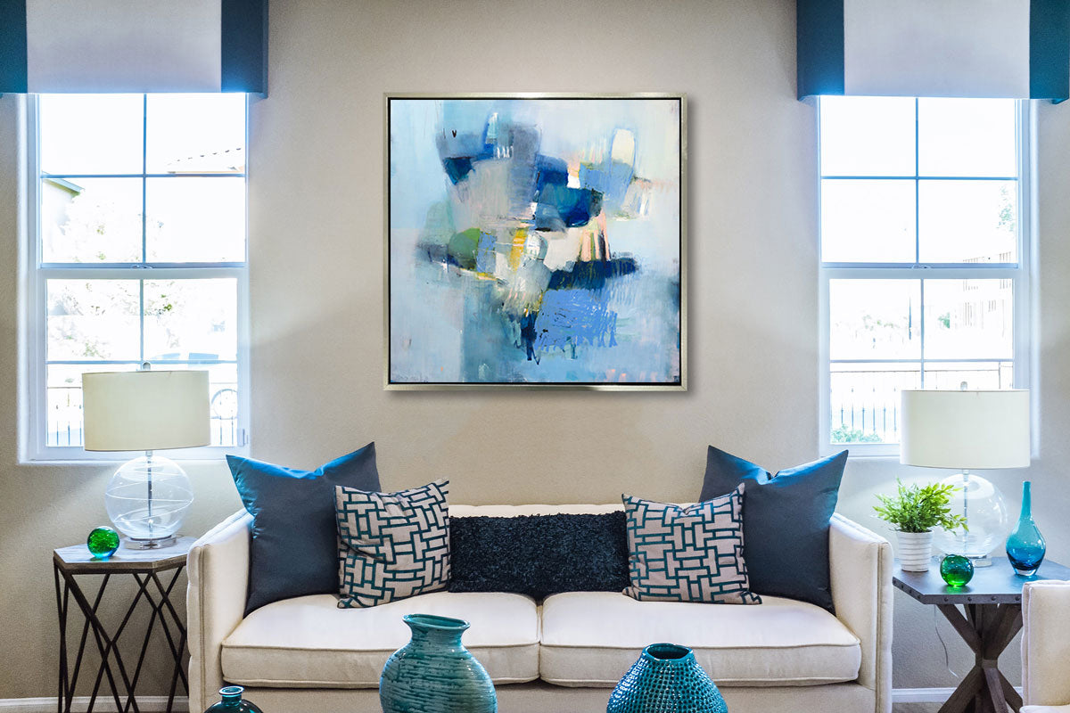 A blue abstract print in a silver floater frame hangs on a white wall above a cream sofa with blue pillows, two side tables with matching lamps and several blue vases.