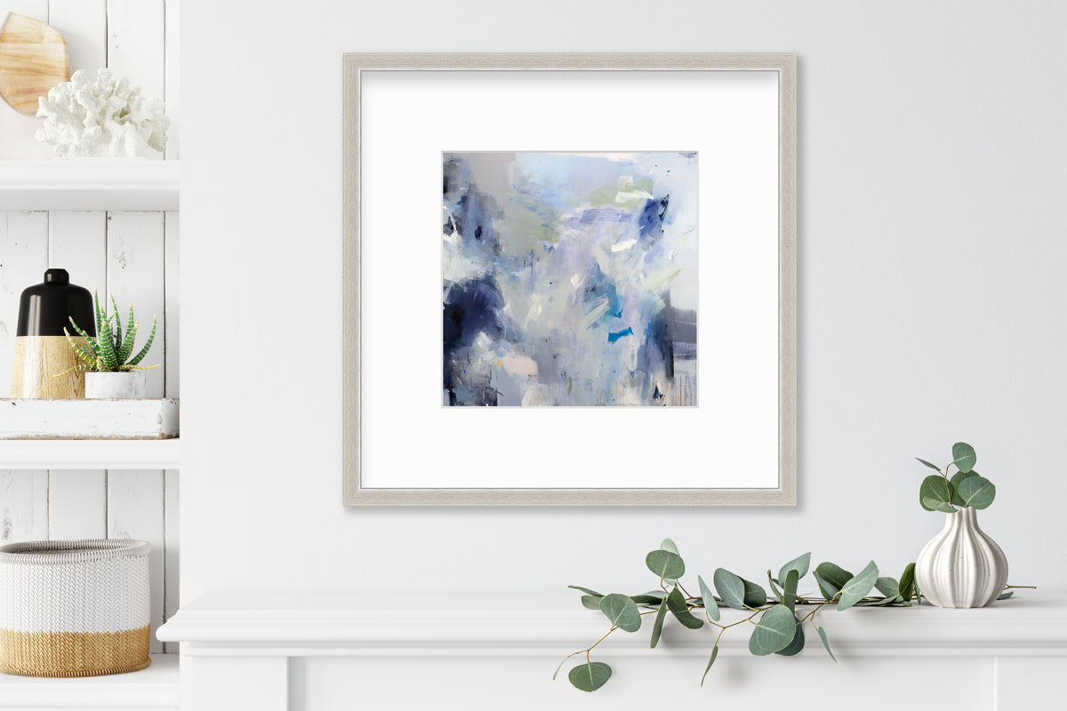 A blue, white, lavender and dark blue abstract print in a silver frame with a mat hangs on a white wall above a white shelf with a eucalyptus filled white vase and next to a built in bookshelf with decorative accessories.