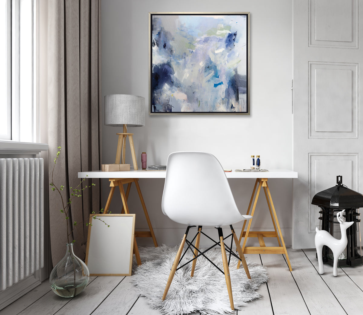 A blue, white, lavender and dark blue abstract print in a silver floater frame hangs on a white wall above a white desk with a natural wood tripod lamp, a white chair, a white faux fur rug and a glass vase on the floor with some greenery stems.