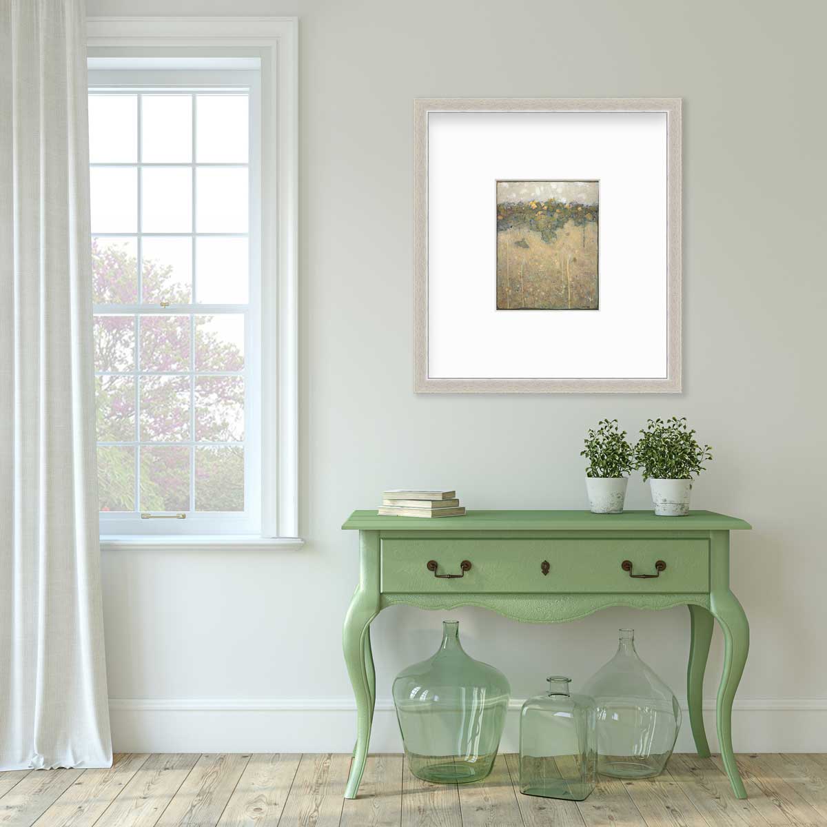 A muted yellow, green and beige abstract landscape print in a silver frame with a mat hangs on a white wall above a green console table with a stack of books, two potted plants and three green glass bottles.