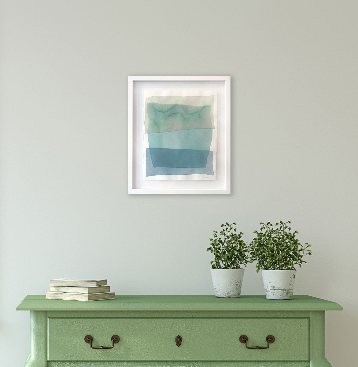 A pale green, teal and blue overlapping shapes painting by Nealy Hauschildt in a white frame floating above a white matte on a white wall above a green console table with a stack of books and two potted plants.