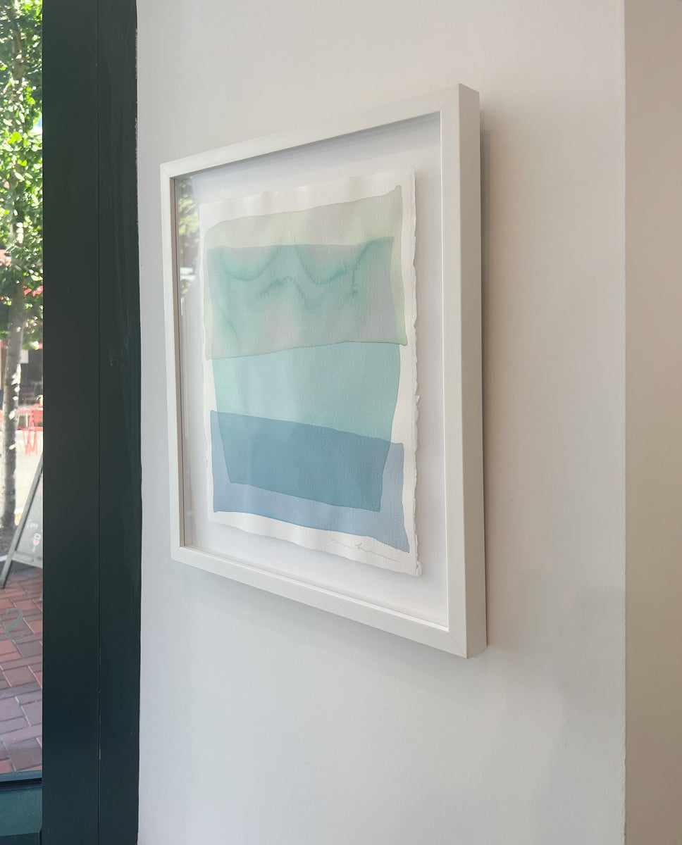 A right side-angle view of a pale green, teal and blue overlapping shapes painting by Nealy Hauschildt in a white frame floating above a white matte on a white wall.