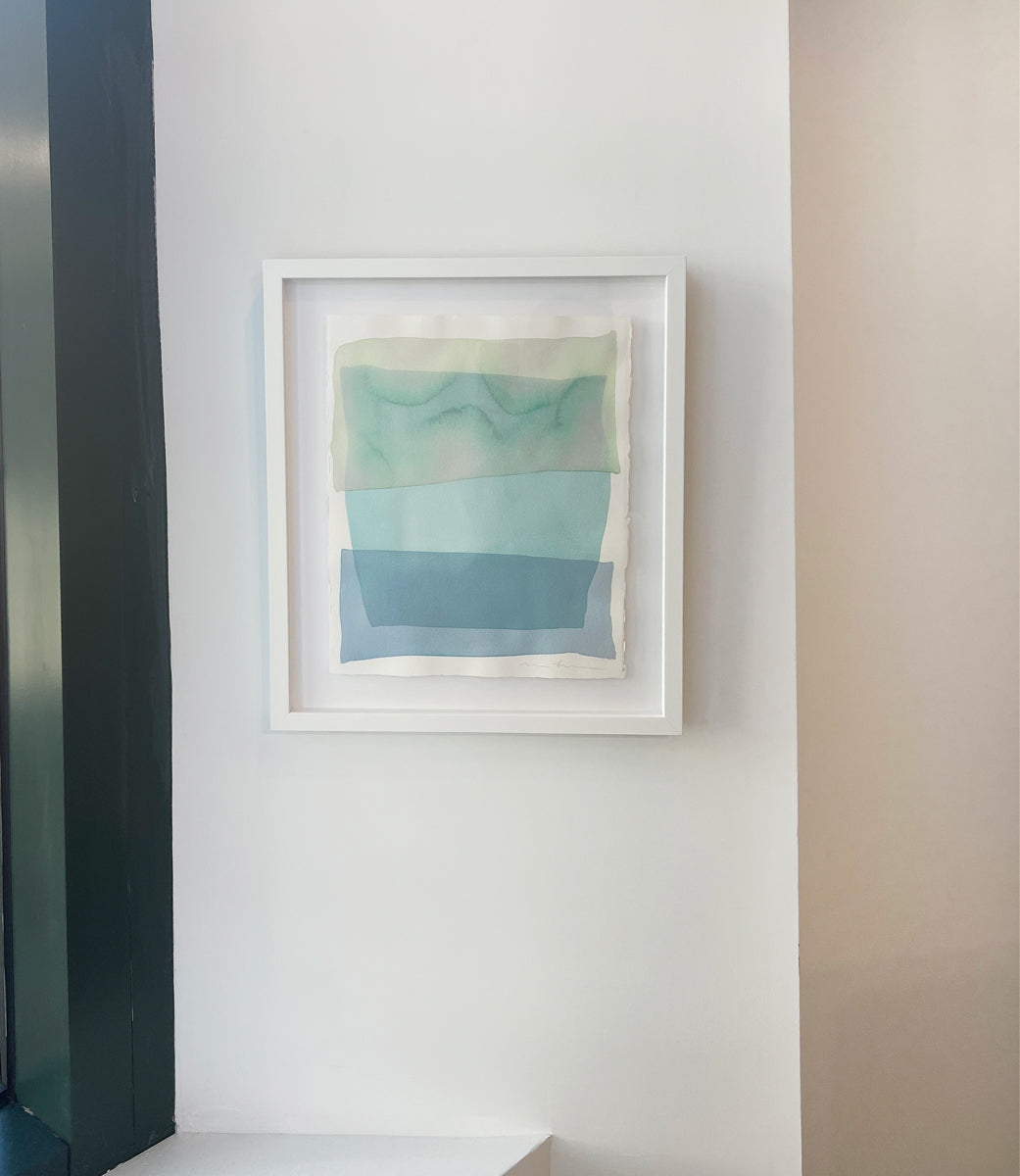 A pale green, teal and blue overlapping shapes painting by Nealy Hauschildt in a white frame floating above a white matte on a white wall.