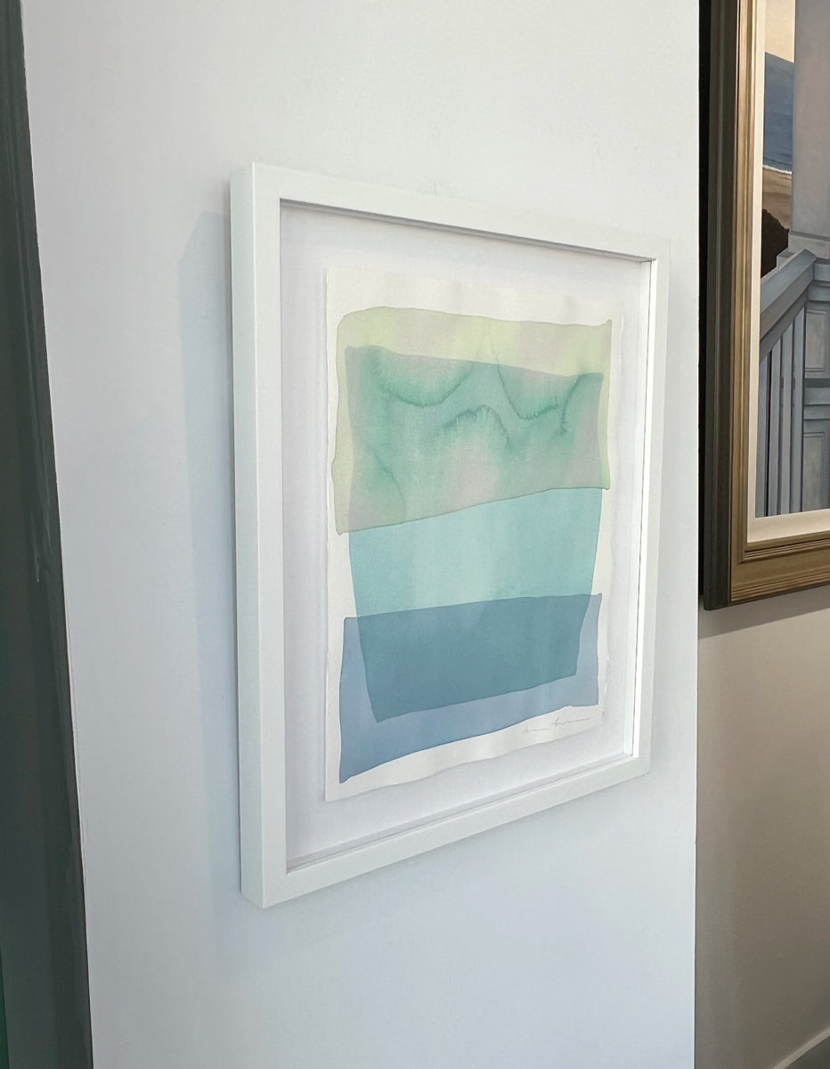 A left side-angle view of a pale green, teal and blue overlapping shapes painting by Nealy Hauschildt in a white frame floating above a white matte on a white wall.