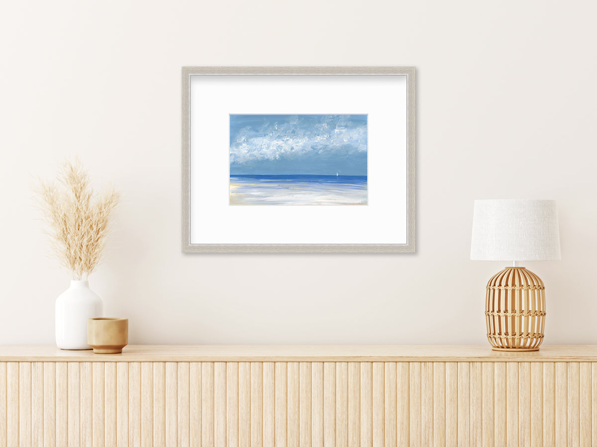 A blue, white and light blue abstract seascape in a silver frame with a mat hangs on a white wall above a natural wood surface with a white vase with wheat stems and a natural wood table lamp.