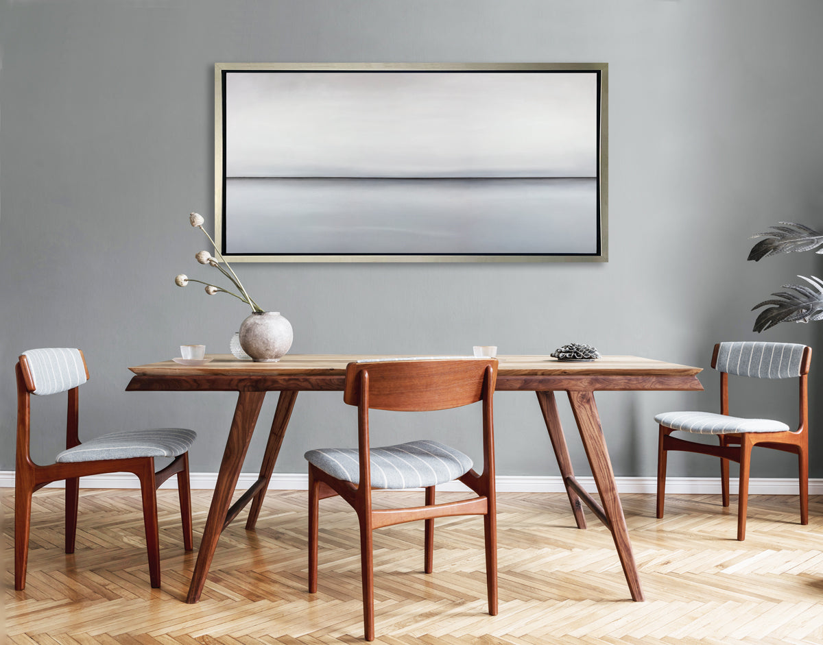 A light grey toned abstract landscape print in a silver floater frame hangs on a grey wall above a wood table and three wood chairs with grey and white striped cushioning.