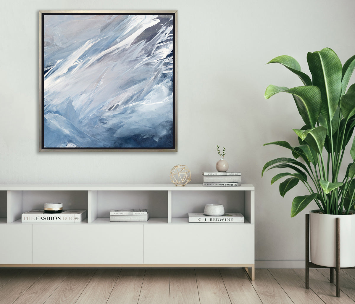 A blue, grey, white and charcoal abstract print in a silver floater frame hangs on a white wall above a white shelf credenza with stacks of books, a gold wire sphere and a floor potted plant.