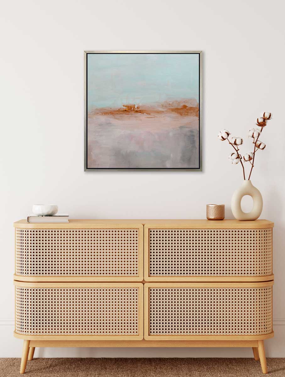A teal, copper and dusty pink abstract print in a silver floater frame hangs on a white wall above a wood console cabinet with a white dish on top of a book, a copper small vase and a white modern vase with cotton stems.