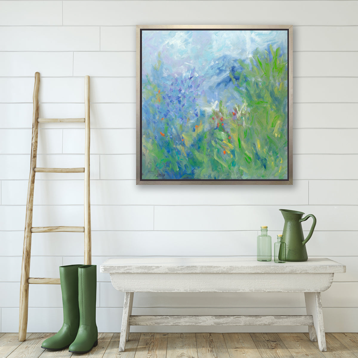 A blue, green, yellow and white abstract floral print in a silver floater frame hangs on a white wall above a whitewashed bench with a green pitcher and green bottles, a pair of green rubber boots and a ladder against the wall.