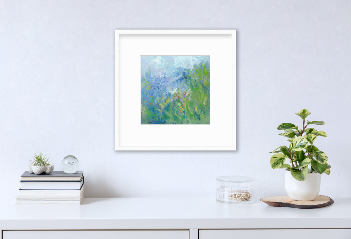 A blue, green, yellow and white abstract floral print in a white frame with a mat hangs on a white wall above a white surface with a stack of books, a round clear container with gold accessories and a potted plant.