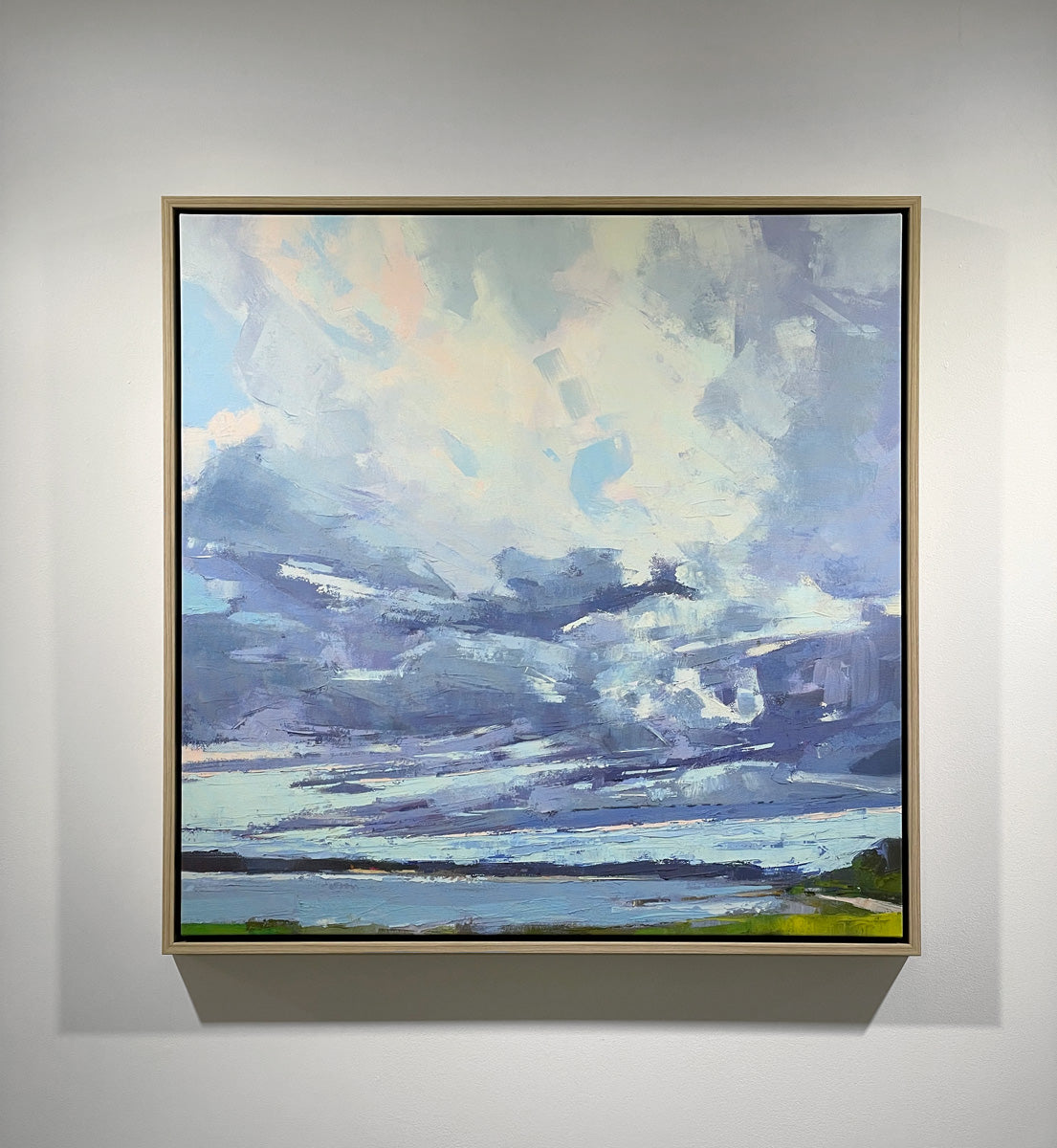 A blue, white and green abstract landscape print in a white oak floater frame hangs on a white wall.