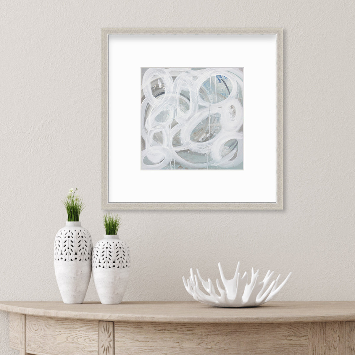 A white, light teal and grey abstract print in a silver frame with a mat hangs on a white wall above a natural wood console table with two white vases and a white decorative bowl.