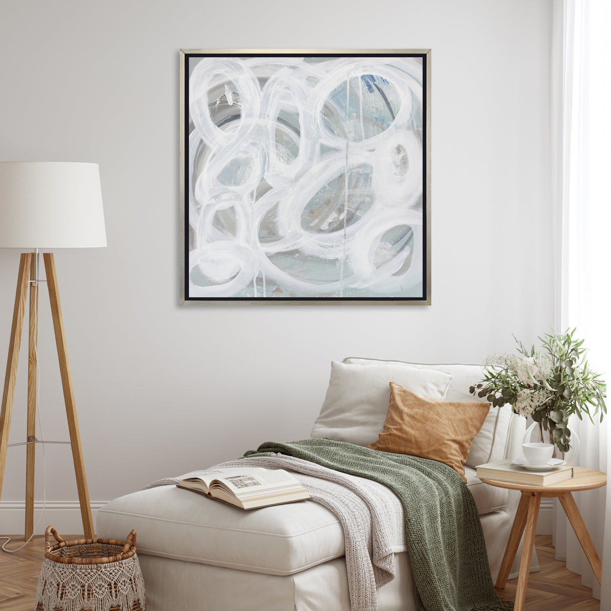 A white, light teal and grey abstract print in a silver floater frame hangs on a white wall above a white chaise lounge with pillows and green throw blanket, a natural wood side table with a book and greenery filled vase and a tripod floor lamp.