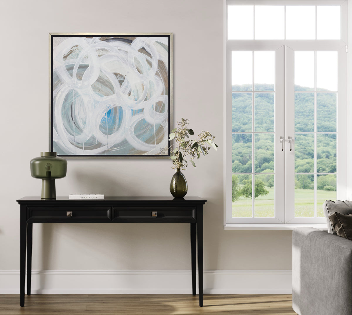 A white, light teal, blue and beige abstract print in a silver floater frame hangs on a white wall above a black console table with a green modern lamp, a deep green vase with white flowers and next to a large window showing a lush mountainside.