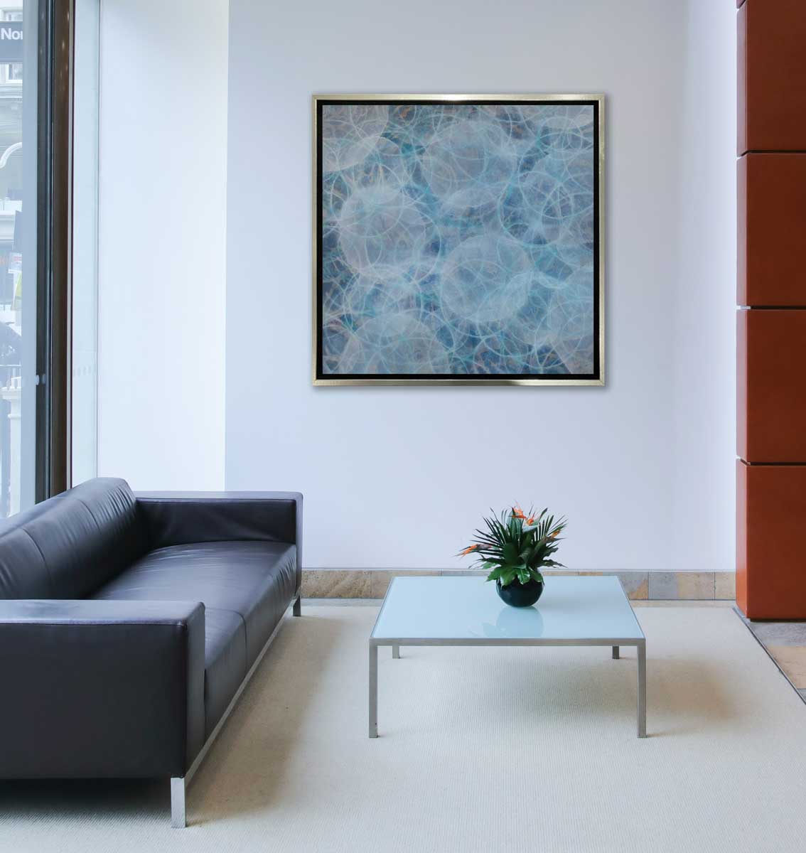 A blue and teal abstract overlapping circle print in a silver floater frame hangs on a white wall above a low back charcoal sofa and a square coffee table with a flower arrangement in the middle.