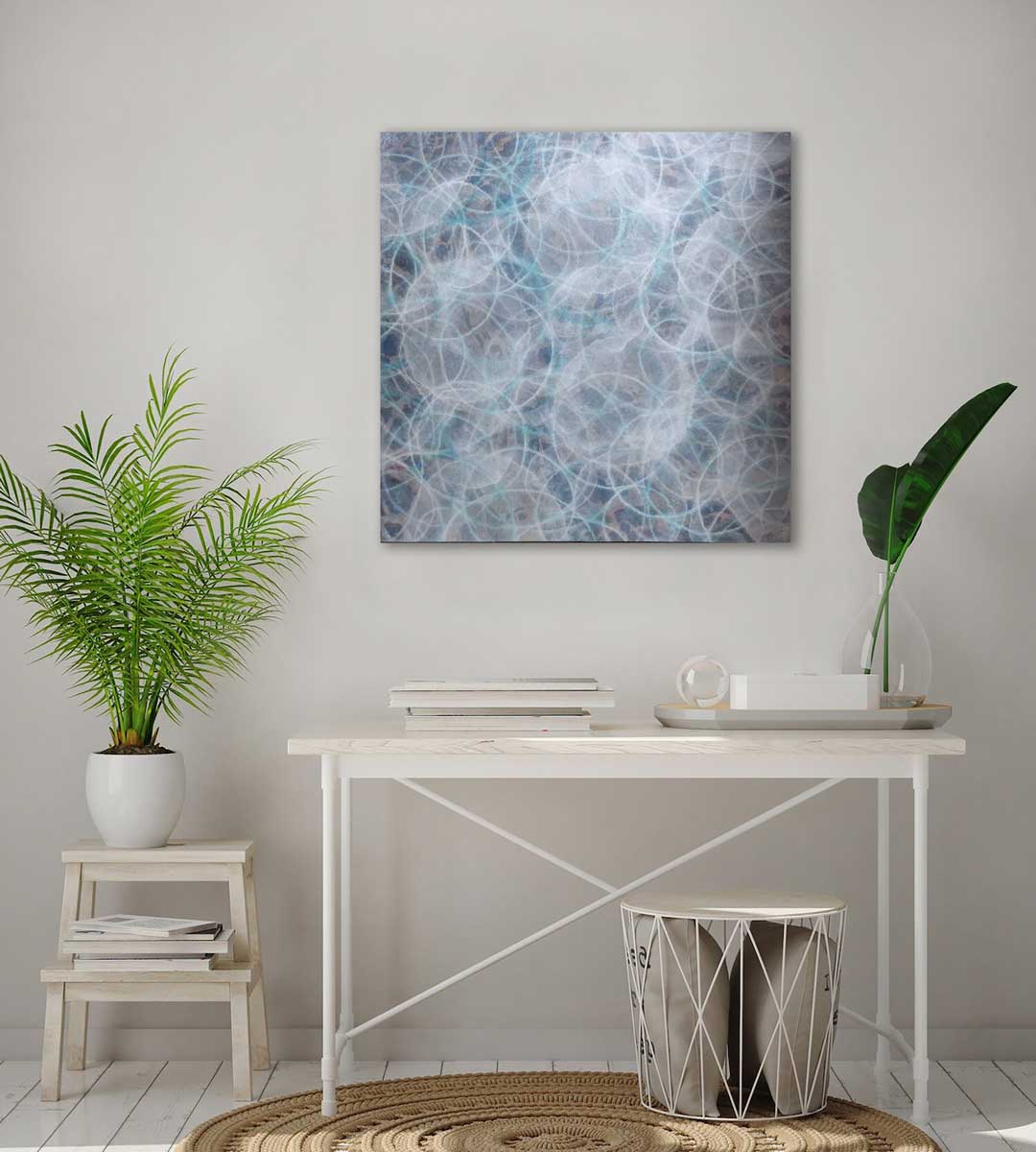 A blue, white and teal overlapping circles painting hangs on a white wall above a white desk with accessories, a small step stool with a potted plant and a basket on the floor holding pillows.