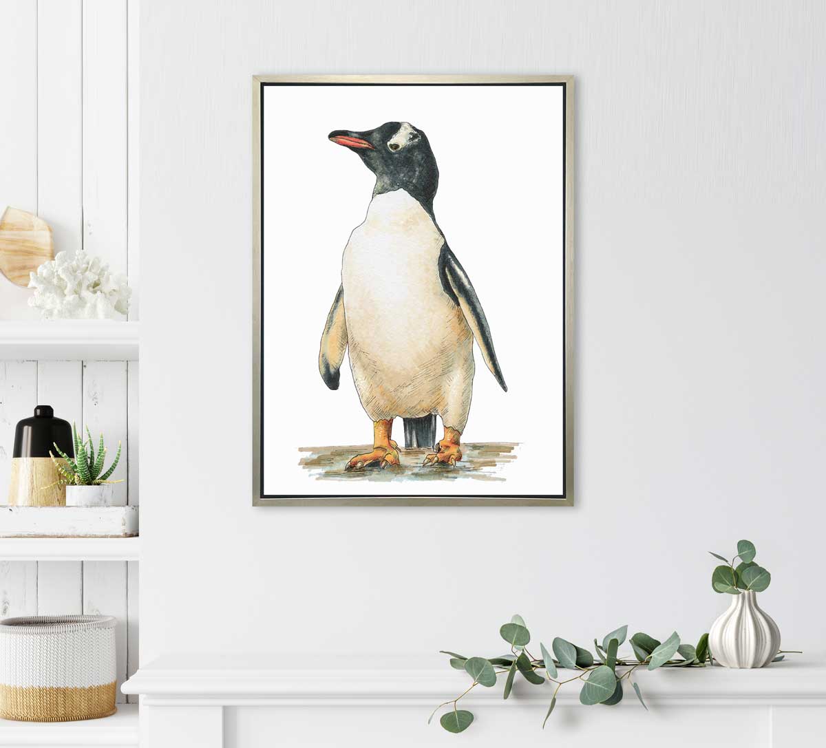 A penguin print in a silver floater frame hangs on a white wall above a small plant in a vase and to the right of white floating shelves holding a variety of decor. 