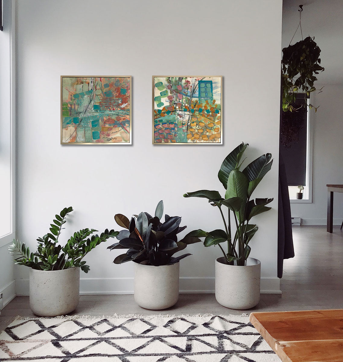 Two abstract encaustic paintings framed in gold frames hang on a grey wall above three large potted plants and a black and white boho carpet. 