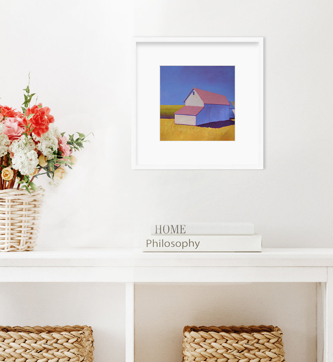 A white barn with a pink roof and yellow grass print in a white frame with a mat hangs on a white wall above a white console with wicker baskets underneath and books stacked on top. 