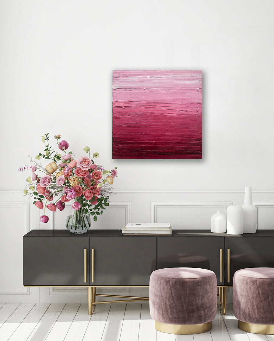 A pink, raspberry and white toned textured painting by Teodora Guererra hangs on a white wall above a dark wood credenza with white vases and a clear vase with red flowers.
