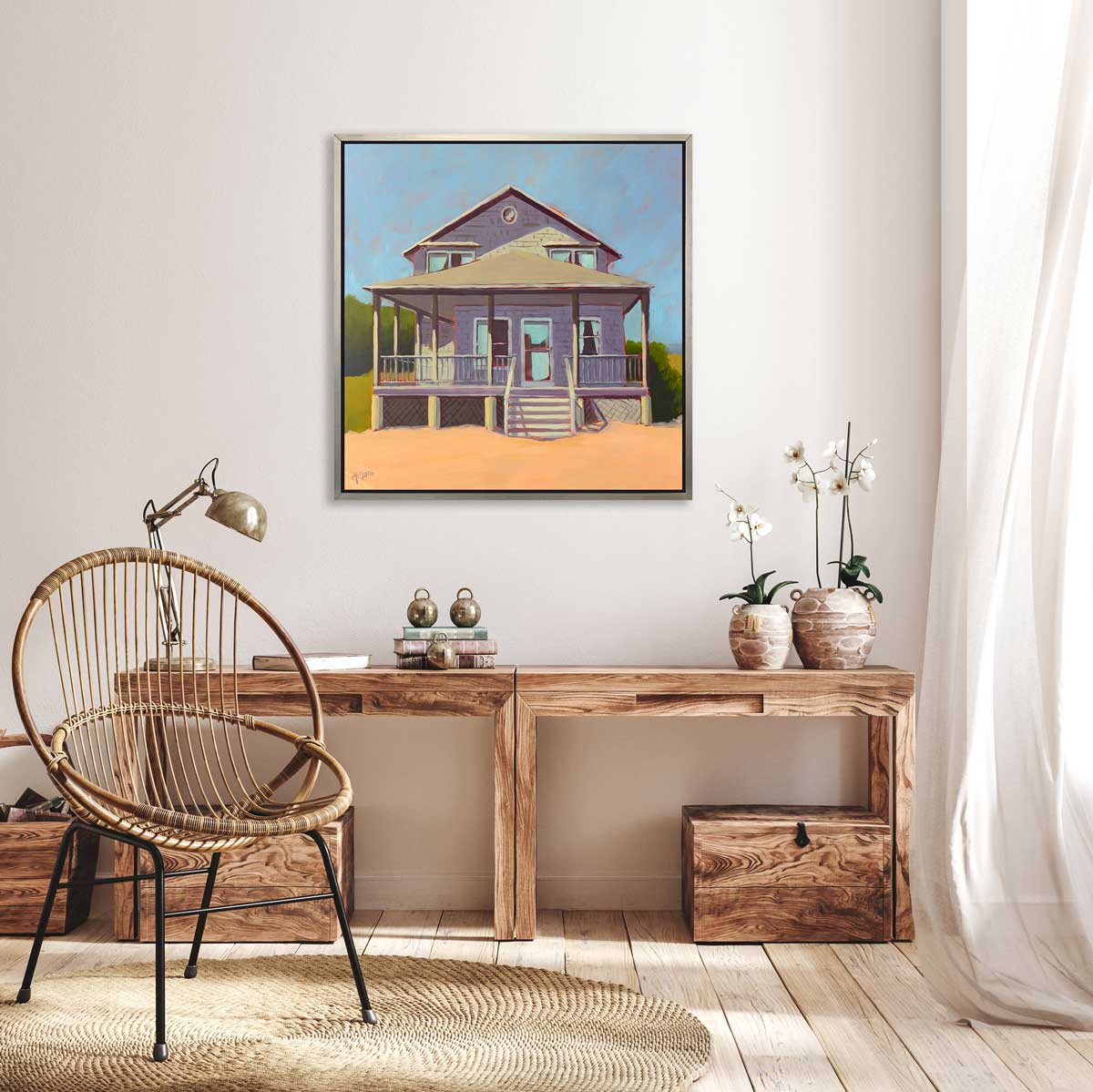 A blue, lavender and peach contemporary beach house print in a silver floater frame hangs on a white wall above a natural wood desk with a stack of books, a chrome desk lamp and two potted white orchids.