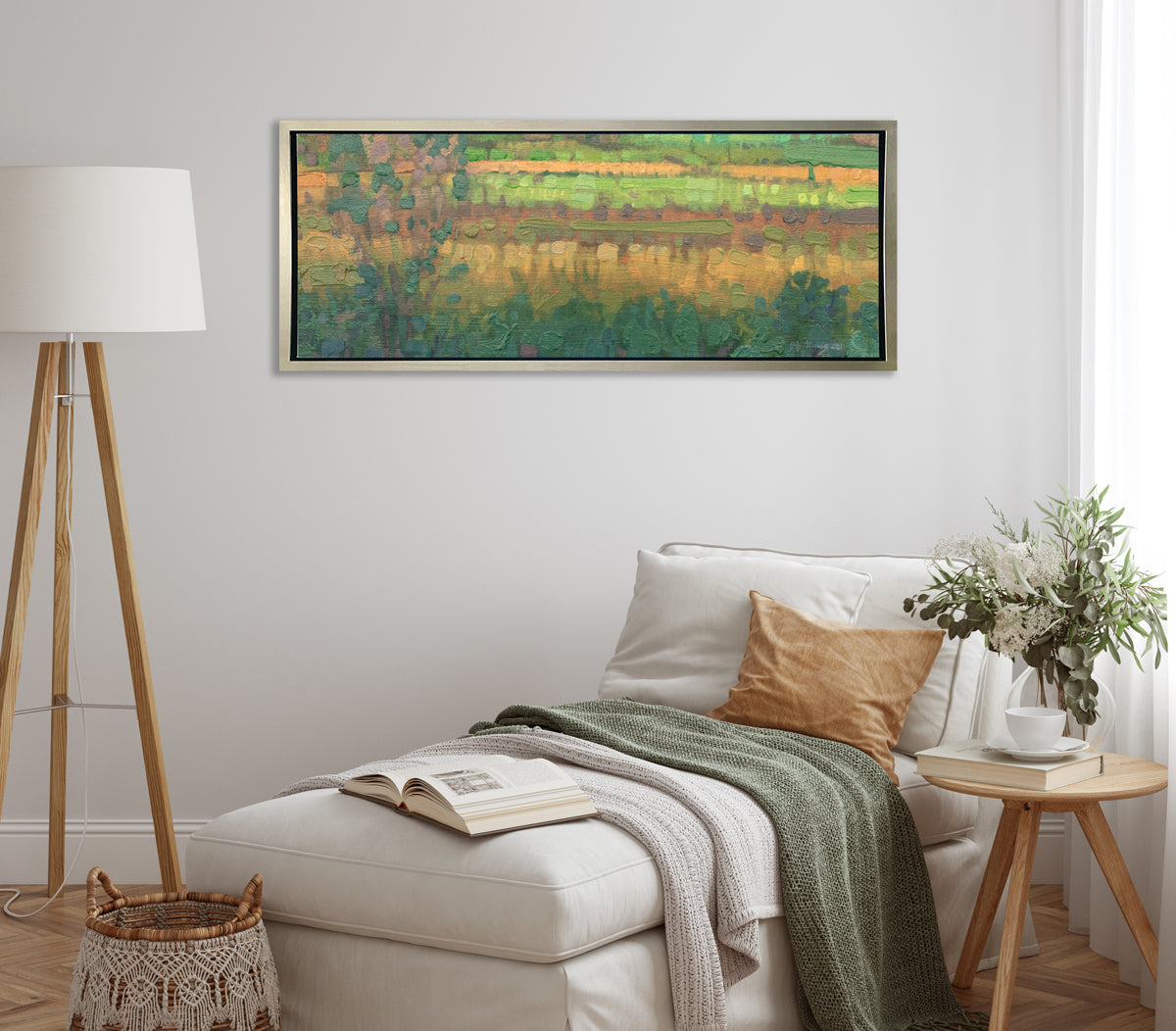 A green, yellow and deep orange abstract landscape print in a silver floater frame hangs on a white wall above a white lounge chair with pillows, a green throw blanket, an open book and a wood side table with a glass vase with a floral arrangement.