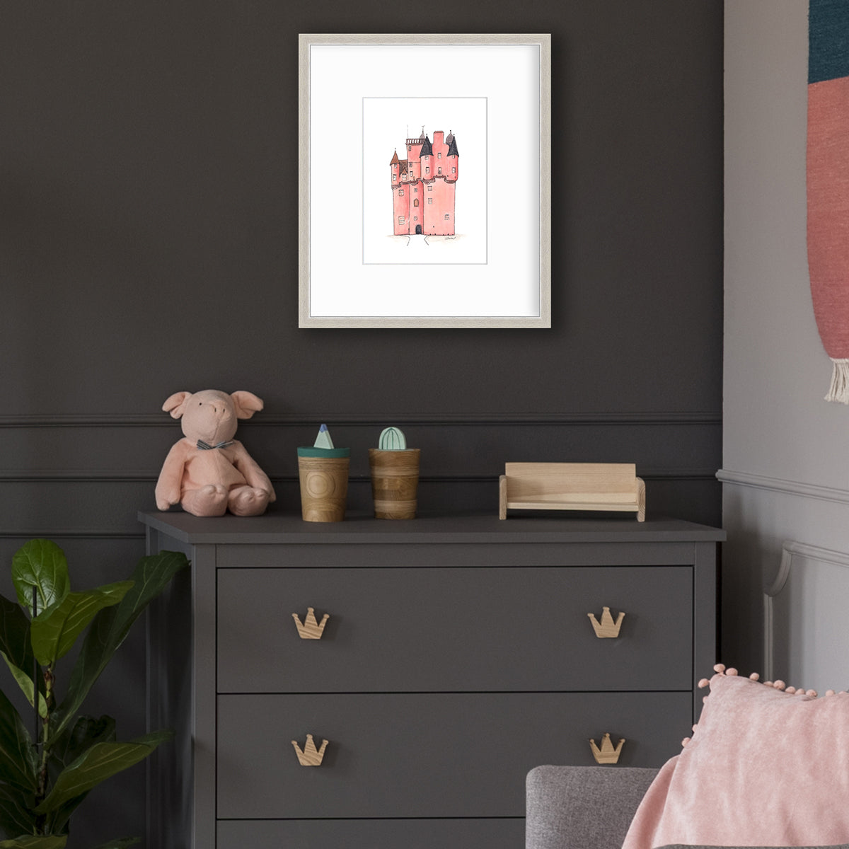 A pink and grey castle print in a silver frame with a mat hangs on a dark grey wall above a dark grey dresser with wood crown drawer pulls, a pig stuffed animal, two wood decorative accessories and a small wood bench decoration.